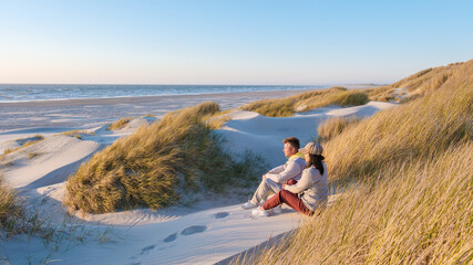As the sun sets over the serene beach in Blokhus Strand,Denmark, a couple sits together on the warm sand dunes, surrounded by lush grass, savoring the tranquil moment.