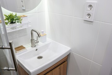 clean, bright bathroom corner features a white sink with a modern faucet, wood-look vanity, and white tiled walls.  Accessories include a plant, soap dispenser, and shelving