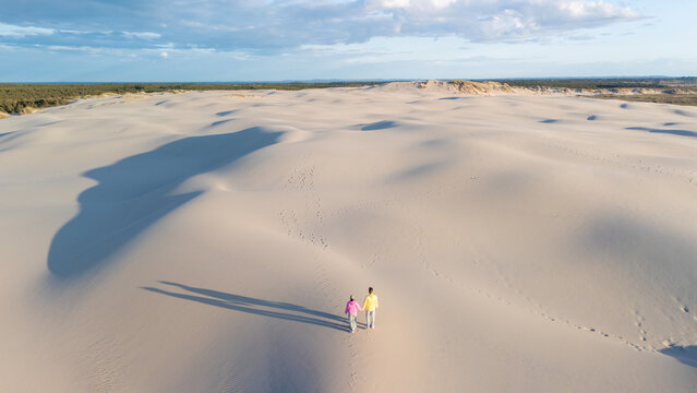 Two individuals stroll hand in hand across the expansive, undulating white sand dunes of Denmark. The warm golden light of sunset casts long shadows, Rabjerg Mile Dunes, Denmark.
