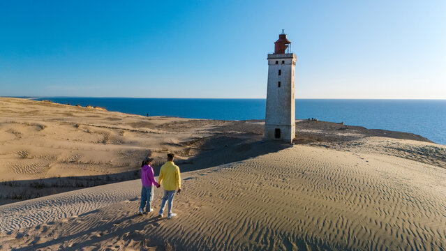 A couple stands hand in hand on the sandy dunes near Rubjerg Knude lighthouse, overlooking the breathtaking coastline of Denmark during a golden sunset. - Powered by Adobe