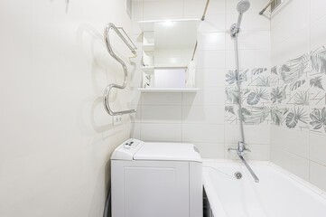 Bright bathroom featuring a white washing machine, tub, tiled walls with leaf-patterned accents, and a mirrored cabinet