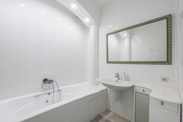 Bright bathroom with white tiles, tub, sink, and framed mirror. Spotlights illuminate the room