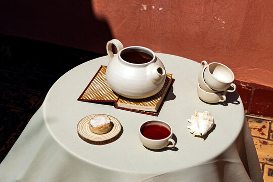 Ceramic teapot and cups on a sunlit terrace table with a shell and book