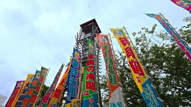 Vibrant, colorful flags (nobori) celebrating sumo wrestlers stand tall against the sky near a traditional tower, indicating a festive sumo atmosphere.
