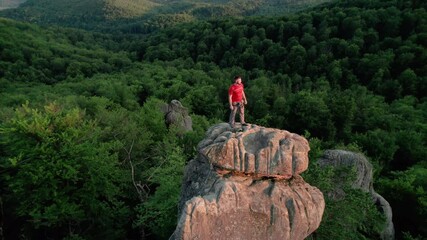 Aerial view of climber standing atop rock formation at Dovbush Rocks in Carpathian mountains, Ukraine. Sun sets, casting warm glow over lush, green forest, distant hills, creating breathtaking view.