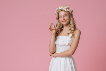 Fototapeta premium Happy girl with flower wreath on her head with perfume bottle in her hand on pink background. Joyful blonde girl with perfumes on pink background.