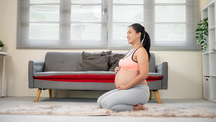 Pregnant asian woman holding her bare belly and sitting kneeling on soft fur carpet in living room.