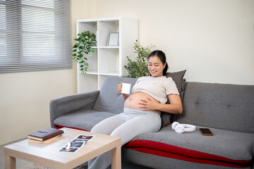 Pregnant asian woman holding a mug as she looking and touching her bare belly while sitting on sofa.