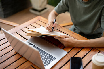 Close up of a hand holding and pointing a pen on notebook with laptop aside sitting at wooden table.