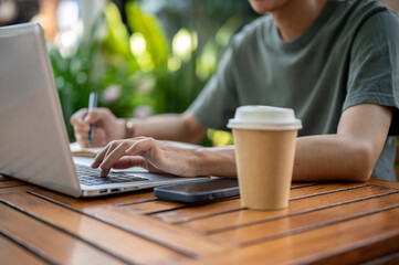 Close up of a hand holding pen and typing on laptop with coffee cup and phone aside on wooden table.