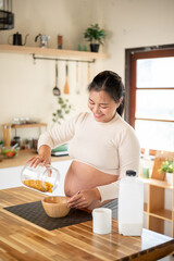 Pregnant asian woman smiling and pouring cereal into a wooden bowl making a meal at kitchen counter.