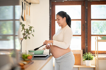 Side view of a pregnant asian woman standing and cooking a meal with a pan on stove in the kitchen.