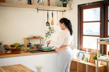 Back view of a pregnant asian woman standing and cooking a meal with a pan on stove in the kitchen.