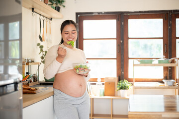 Pregnant asian woman holding a fork enjoy eating vegetable salad from glass bowl standing in kitchen