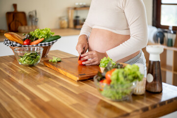 Close up of pregnant asian woman cutting tomato with knife on chopping board making salad in kitchen