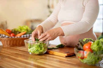 Close up of pregnant woman putting cucumber slices in glass bowl making vegetables salad in kitchen.