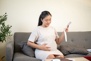 Pregnant asian woman looking at ultrasound pictures and caressing her belly sitting on sofa in house