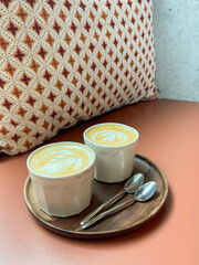 Two steaming cappuccinos with delicate heart-shaped foam designs rest on a rustic wooden tray, surrounded by plush cushion seating in a charming café. Perfect for coffee culture content