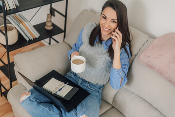 Smiling freelancer talking on phone, drinking coffee and working on laptop at home