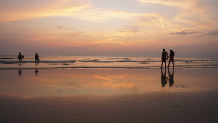 Couple holding hands while walking along a wet beach at sunset, with tourists bathing in the background and the vibrant sky reflecting on the tranquil water surface
