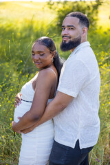 An African American couple enjoys summer outdoors, with the husband lovingly embracing his wifes pregnant belly in a serene, green setting, celebrating their intimate moments and future as a family