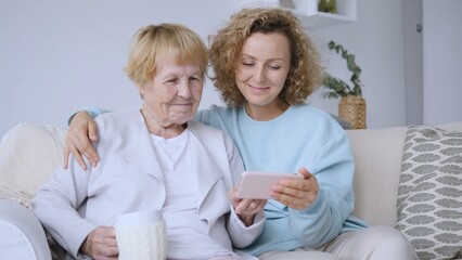 Smiling nurse showing something on a smartphone to a senior woman holding a mug on a sofa, concept of home care and assistance for the elderly