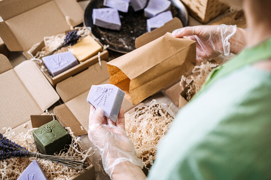 Person wearing gloves places lavender soap into a kraft bag among handmade skincare products. Handmade soap process, lavender skincare, indie soap maker, ethical manufacturing