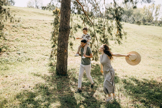 Playful family moment in a sunny park, with mother holding straw hat and father carrying son on shoulders.