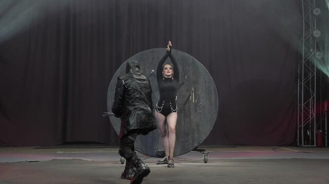 A circus show with throwing knives in the circus arena under the light of colorful spotlights. The assistant girl stands near the wall where a man with a mask on his face throws knives.