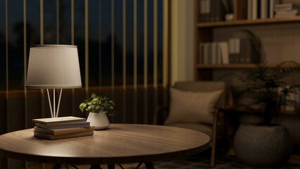 Books and potted plant on wooden round table under lamp light in a living room or a library at night.