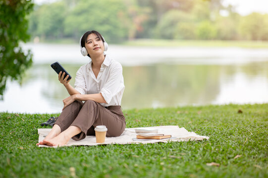 Asian woman wearing headphone listening to music while holding phone sitting at picnic mat on grass.
