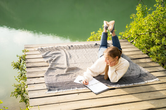 Full body of young woman writing in notebook outdoors near mountain lake enjoying harmony moment alone lying on blanket on wooden pier at sunset, escape from urban hustle, countryside slow living