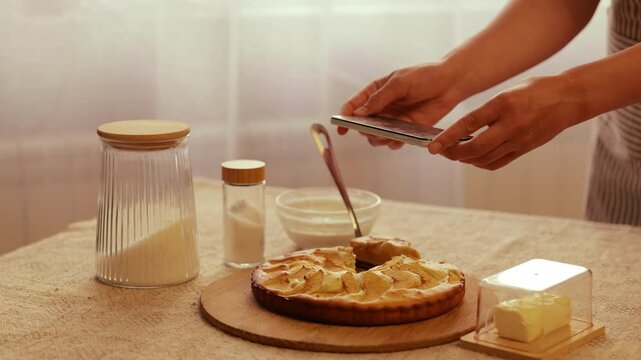 A person uses a smartphone to photograph a freshly baked apple cake on a wooden board, surrounded by baking ingredients in a rustic kitchen setting.