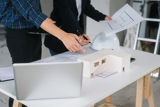 Asian couple reviews architectural plans together at renovation site. They collaborate on ideas to remodel newly purchased second-hand home, standing in front of spiral staircase and unfinished walls