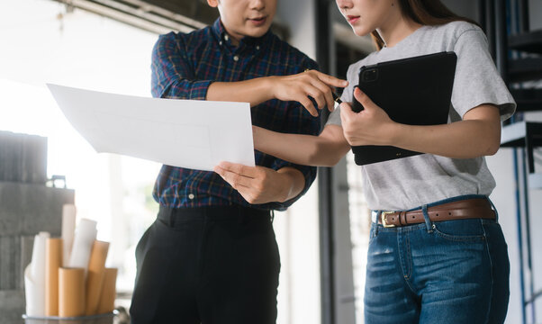Asian couple reviews architectural plans together at renovation site. They collaborate on ideas to remodel newly purchased second-hand home, standing in front of spiral staircase and unfinished walls