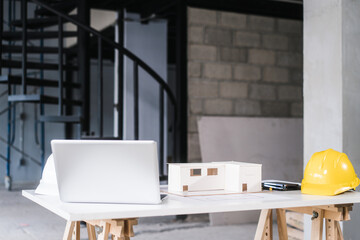 Modern architectural workspace featuring white laptop, scale house model, yellow safety helmet on...