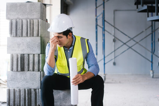 Asian male engineer wearing a hard hat and safety vest sits on concrete blocks, visibly stressed and exhausted, holding building plans amid challenging construction site and unfinished project work.