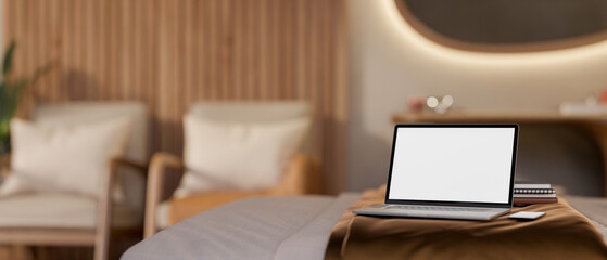 White screen laptop and books on bed in bedroom with led light mirror hanging over wooden armchairs.