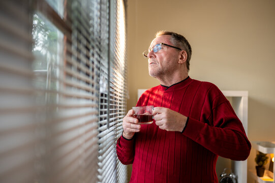 Old white caucasian man wearing glasses holding his coffee cup looking ahead through window blinds.