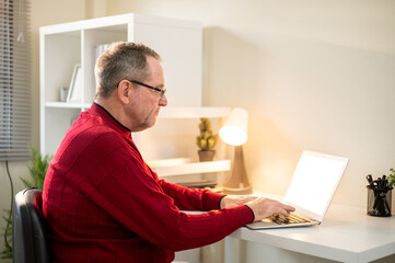 Glasses old white caucasian man typing and looking at laptop with lamp aside on table in living room