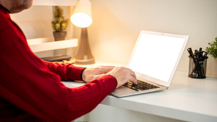 Close up of a man hands typing while looking at white screen laptop on table under lamp light.