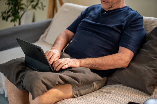 Old white caucasian man typing on tablet keyboard atop pillow on lap while sitting on sofa in house. - Powered by Adobe