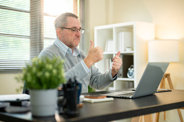 Old white caucasian man wearing glasses making double thumbs up at laptop on office's working table.