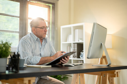 Glasses old white caucasian man with a pen and clipboard looking at computer on office working table