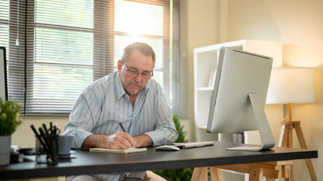 Old white caucasian man wearings glasses writing in notebook with a pen on working table in office.