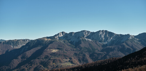 Obraz premium Panoramic view of the Komovi mountains in Montenegro during autumn, with rugged rocky peaks and forested slopes bathed in soft morning light under a clear blue sky.