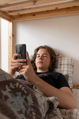 A young man checks his smartphone in bed after waking up in the morning. He is scrolling through social media or news, starting his day connected to the internet and online content.