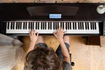 A musician plays a digital piano, seen from a top-down perspective. A smartphone with a learning app rests on the music stand, illustrating online music education or a modern hobby.