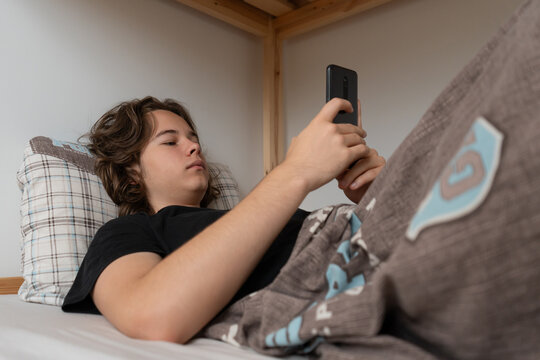 A teenager spends a lazy morning procrastinating in bed, engrossed in his smartphone. Instead of getting up, he browses social media or watches video content, enjoying a slow start to his day.