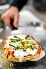 A close-up of a freshly baked pizza being lifted with a spatula. The pizza is topped with melted cheese, herbs, and a drizzle of olive oil, showcasing its appetizing and delicious appearance.